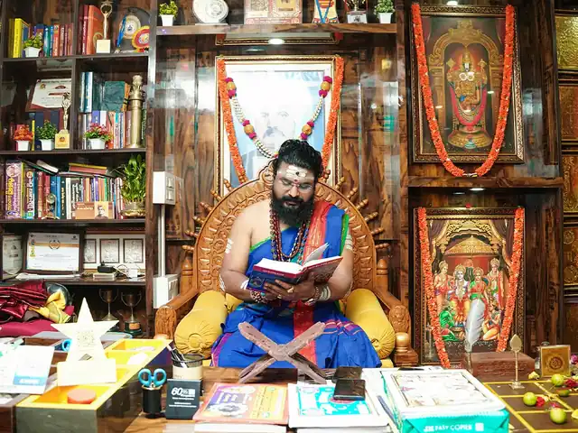 Guruji reading at his desk, with his awards and books on the shelves behind him